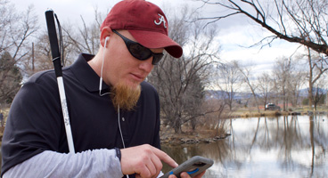 A young blind man wearing ear buds, listens to NFB-NEWSLINE on his phone outside.